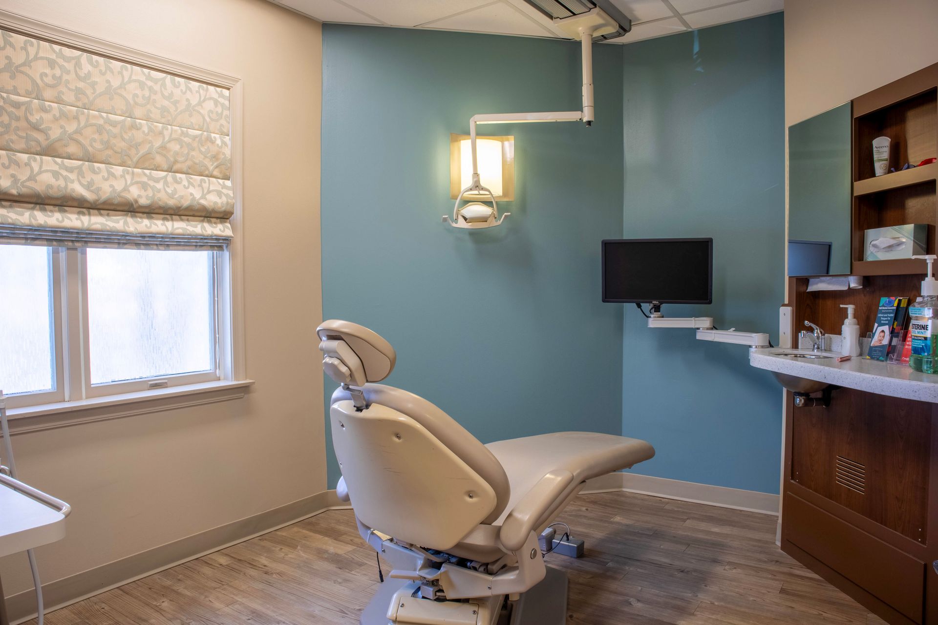 An empty dental examination room featuring a reclining chair, a wall-mounted light, a monitor, and a wooden sink station.