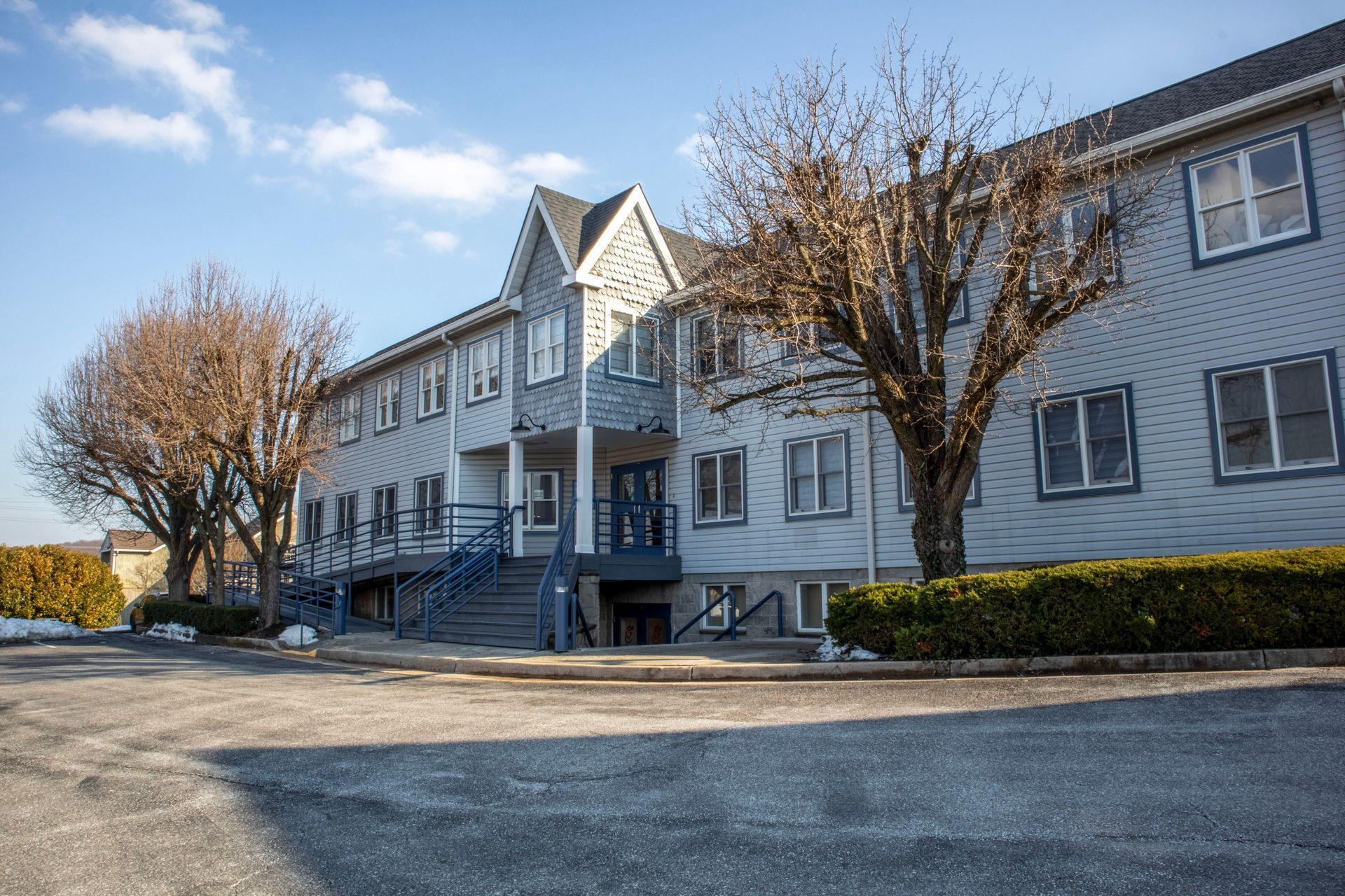 A two-story grey building with a central gabled entrance, stairs, and bare trees under a clear blue sky.