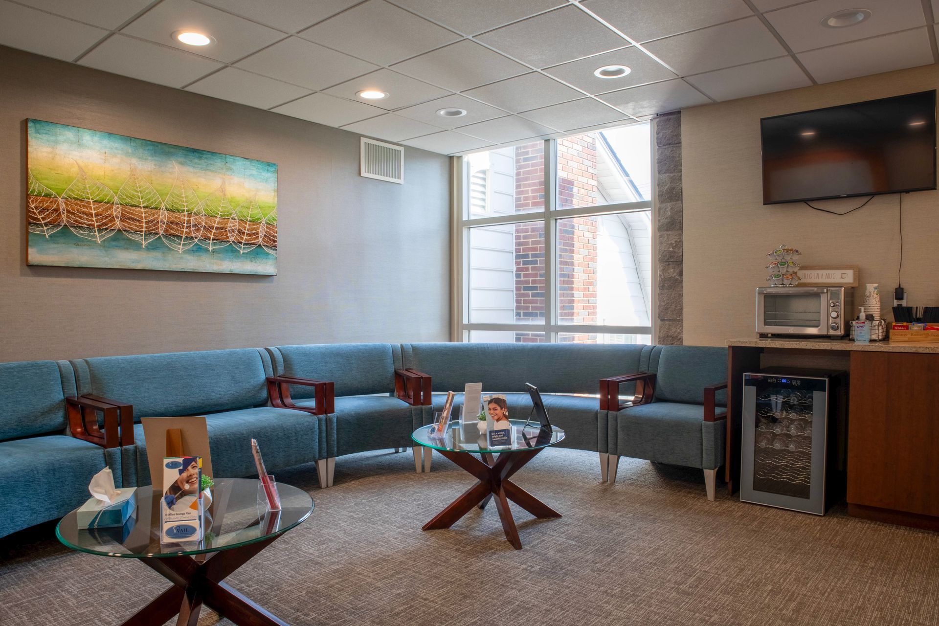 A waiting room with teal sectional seating, two glass coffee tables, a wall painting, a TV, a microwave, and a mini-fridge.