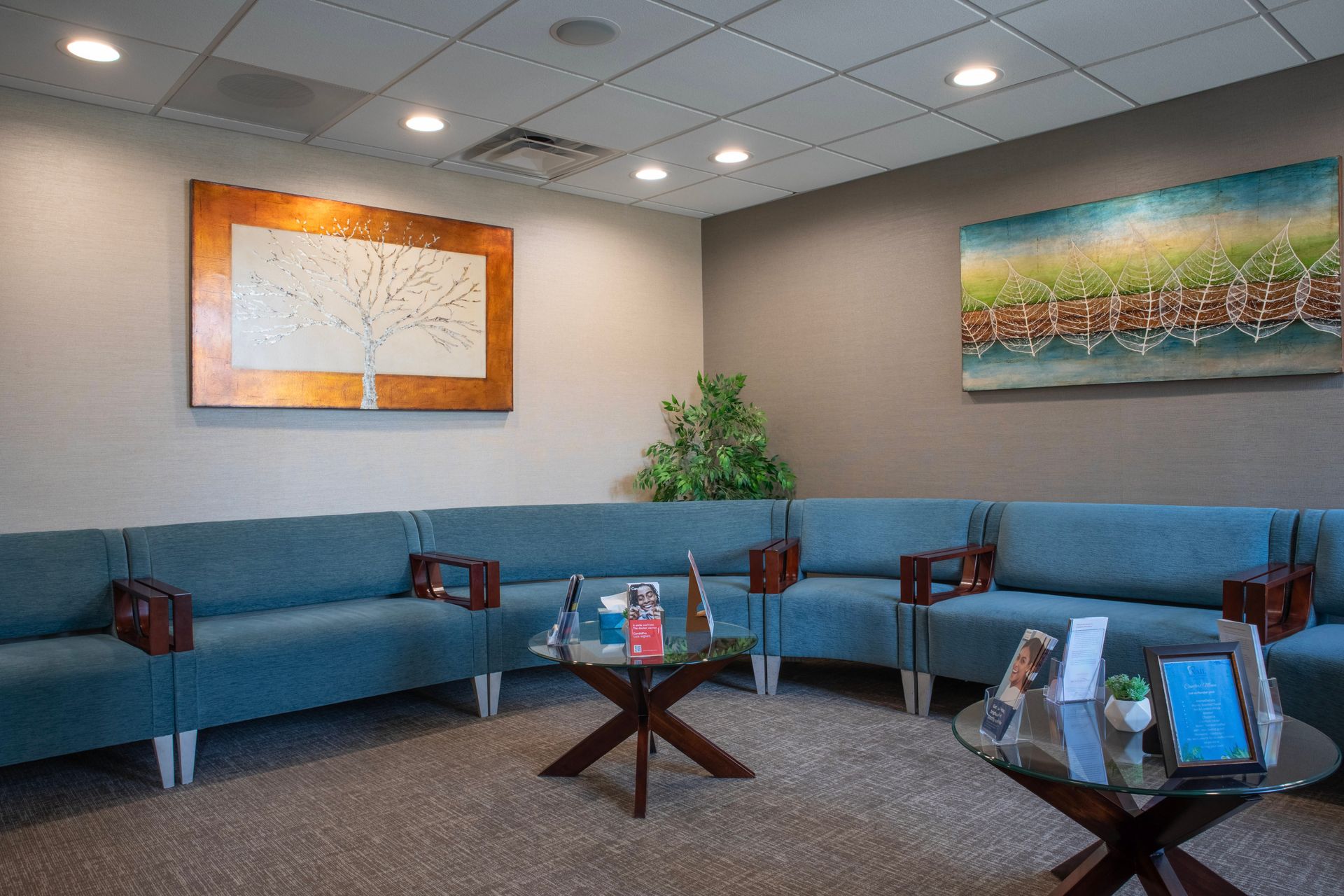 A modern waiting room featuring a blue sectional sofa, a small plant, and two circular coffee tables against gray walls.