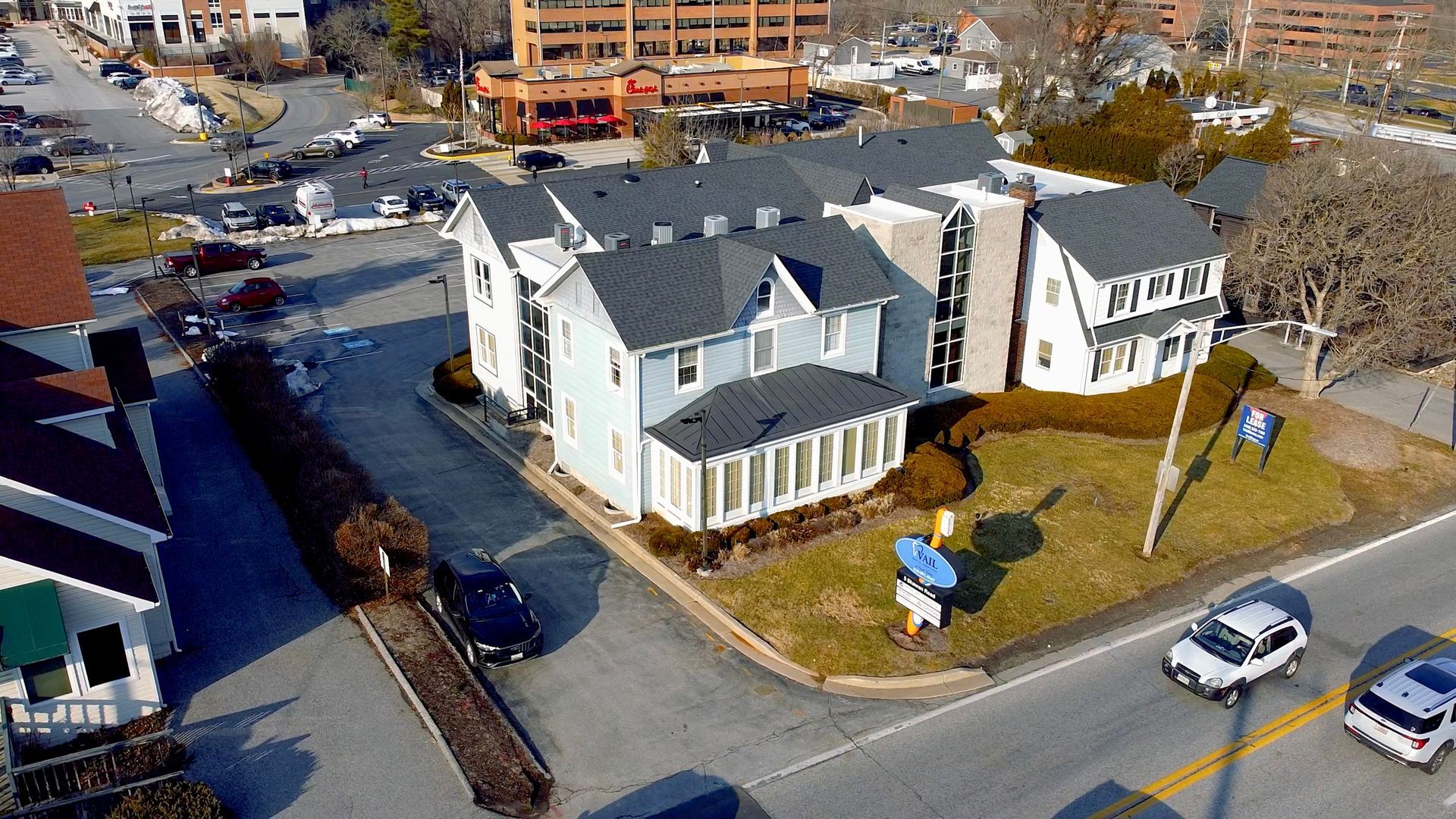 An aerial view of a white, multi-level office building with a dark roof, located on a street with cars in suburban area.