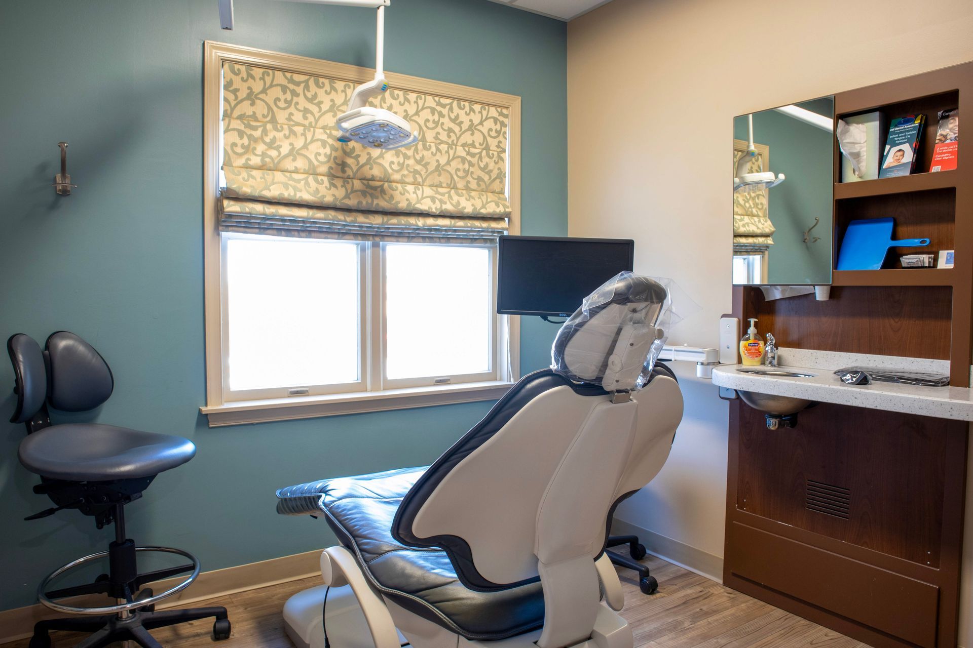 A dental exam room featuring a chair, a practitioner’s stool, a window with a patterned shade, and a small sink cabinet.