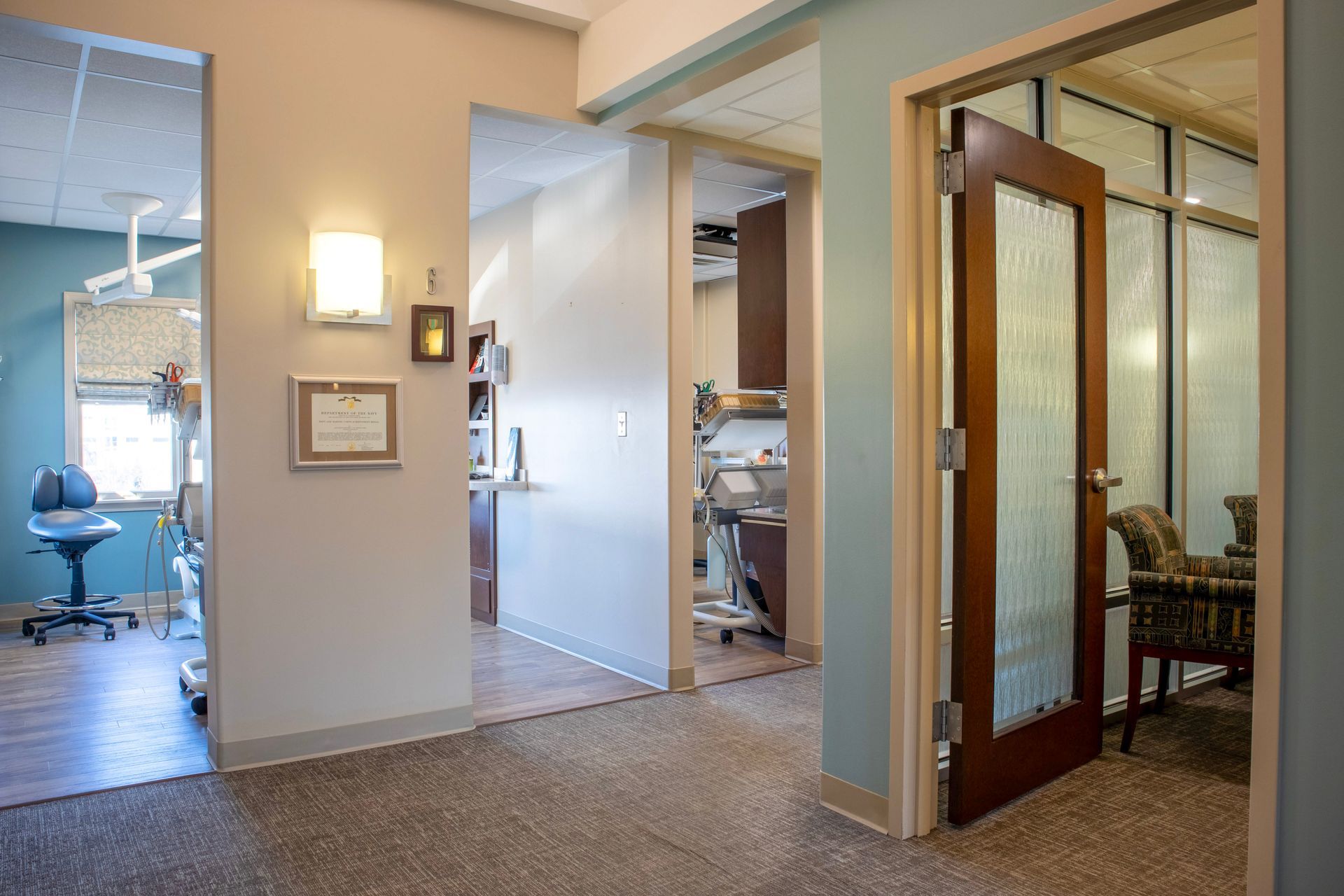 A hallway in a professional office featuring several rooms, glass-paneled doors, neutral carpeting, and blue wall accents.