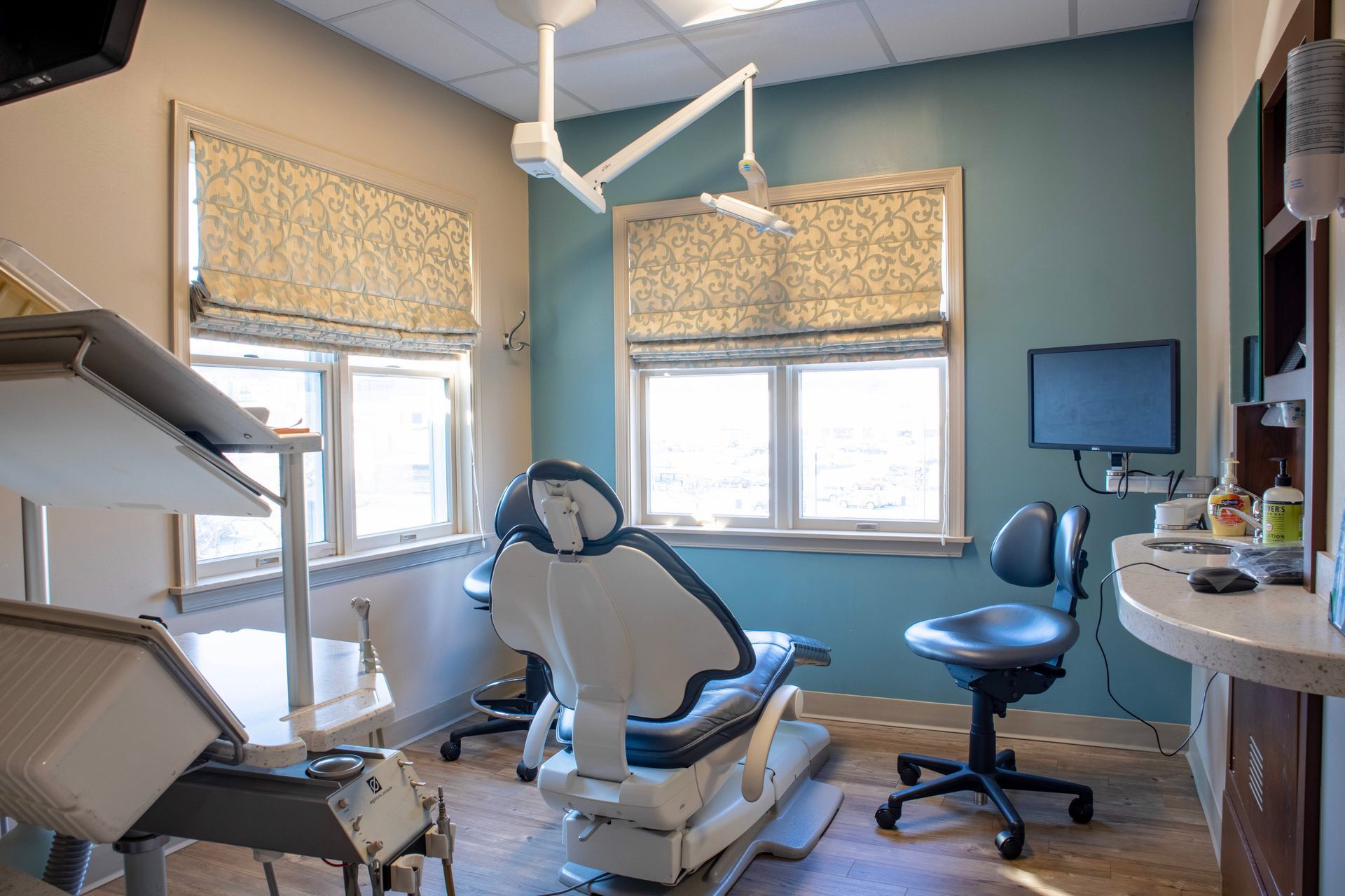 A dental exam room with a central patient chair, an operator stool, dental equipment, and windows with roman shades.