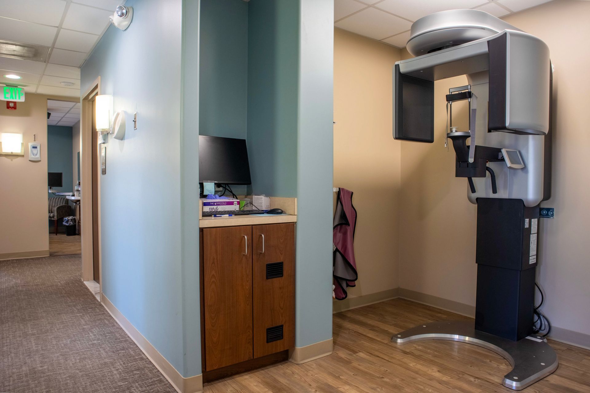A medical dental imaging machine stands in a clinic room next to a wooden cabinet with a computer setup.