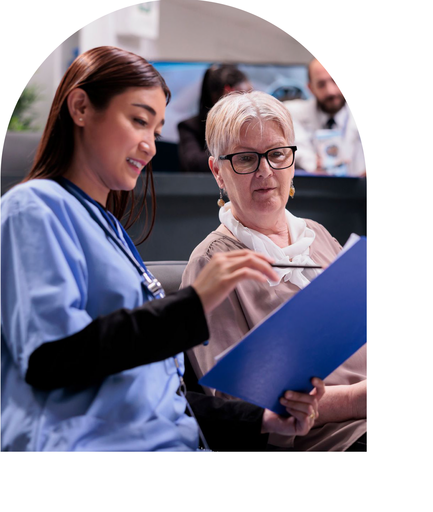 Nurse in blue scrubs points at paperwork with older patient in doctor's office.