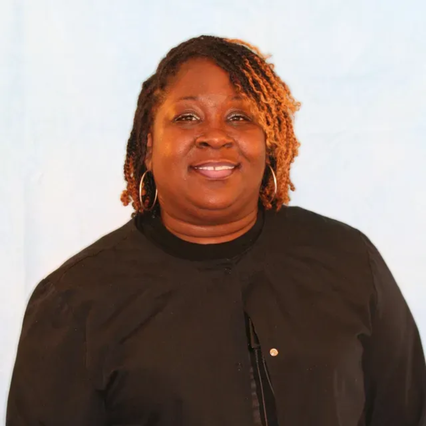 Gina with dreadlocks, wearing a black jacket and hoop earrings, smiling against a light blue background.