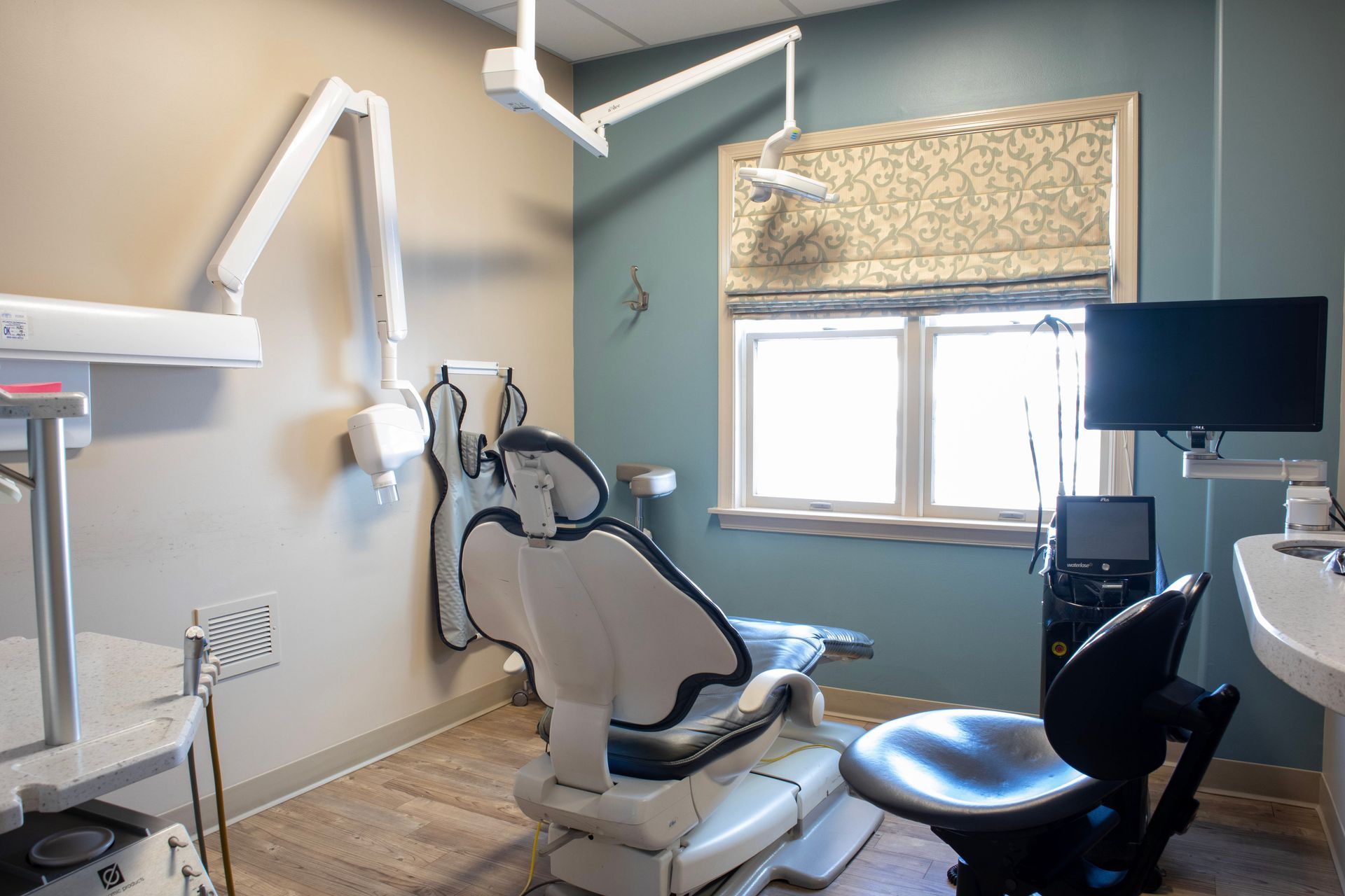 A modern, empty dental operatory with a treatment chair, clinical equipment, a window with a shade, and blue-toned walls.