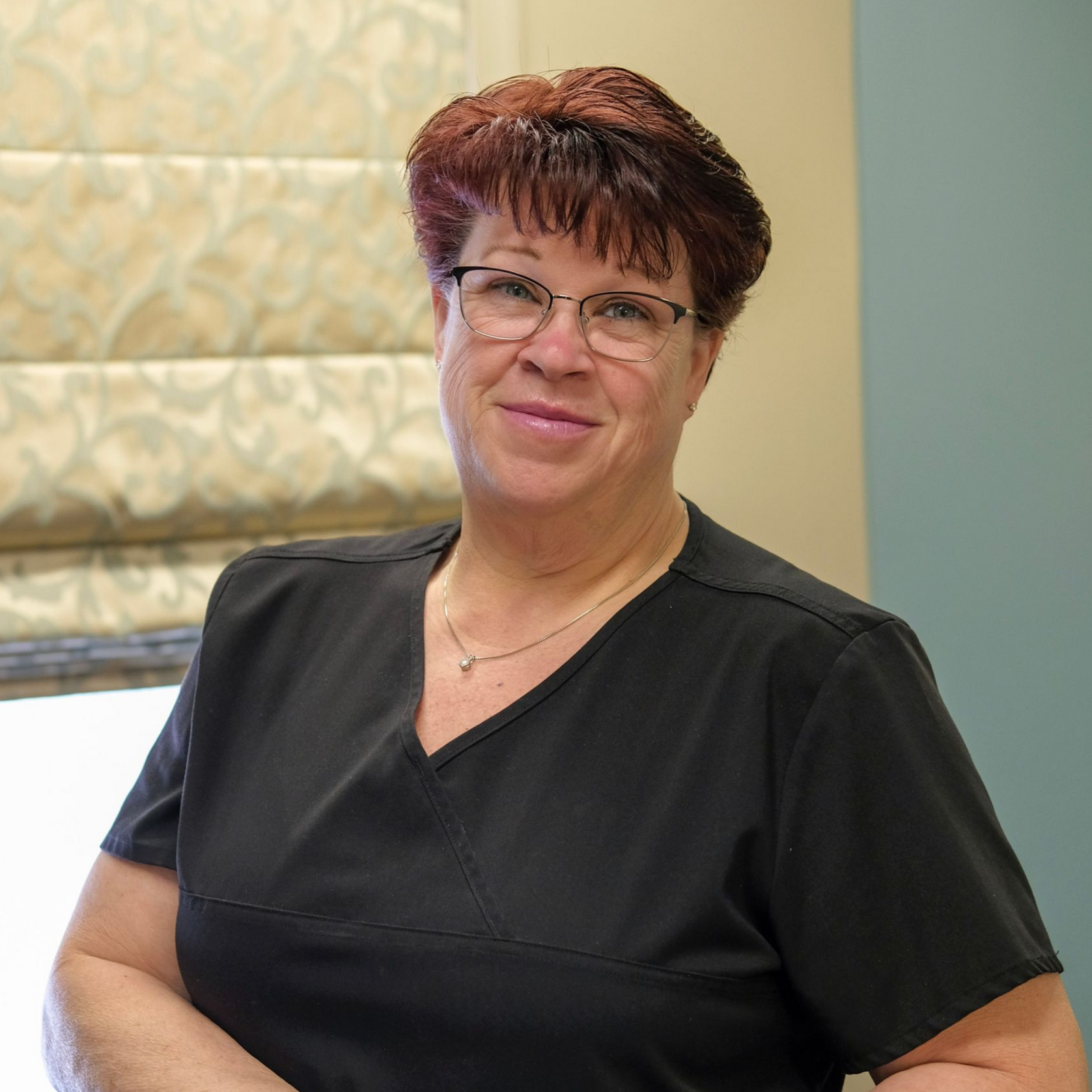 Joanie with glasses and short brown hair, smiling, wearing a black top against a teal background.