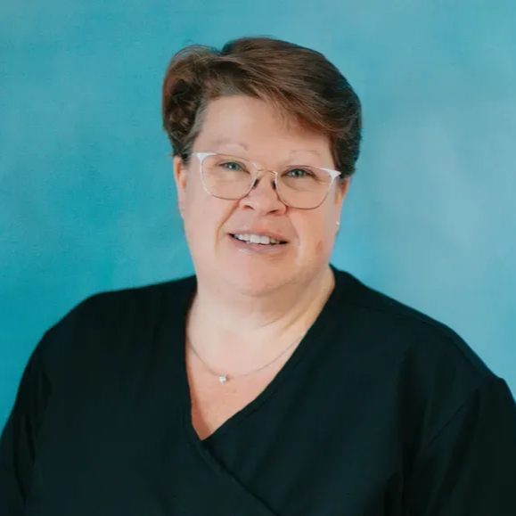 Joanie with glasses and short brown hair, smiling, wearing a black top against a teal background.
