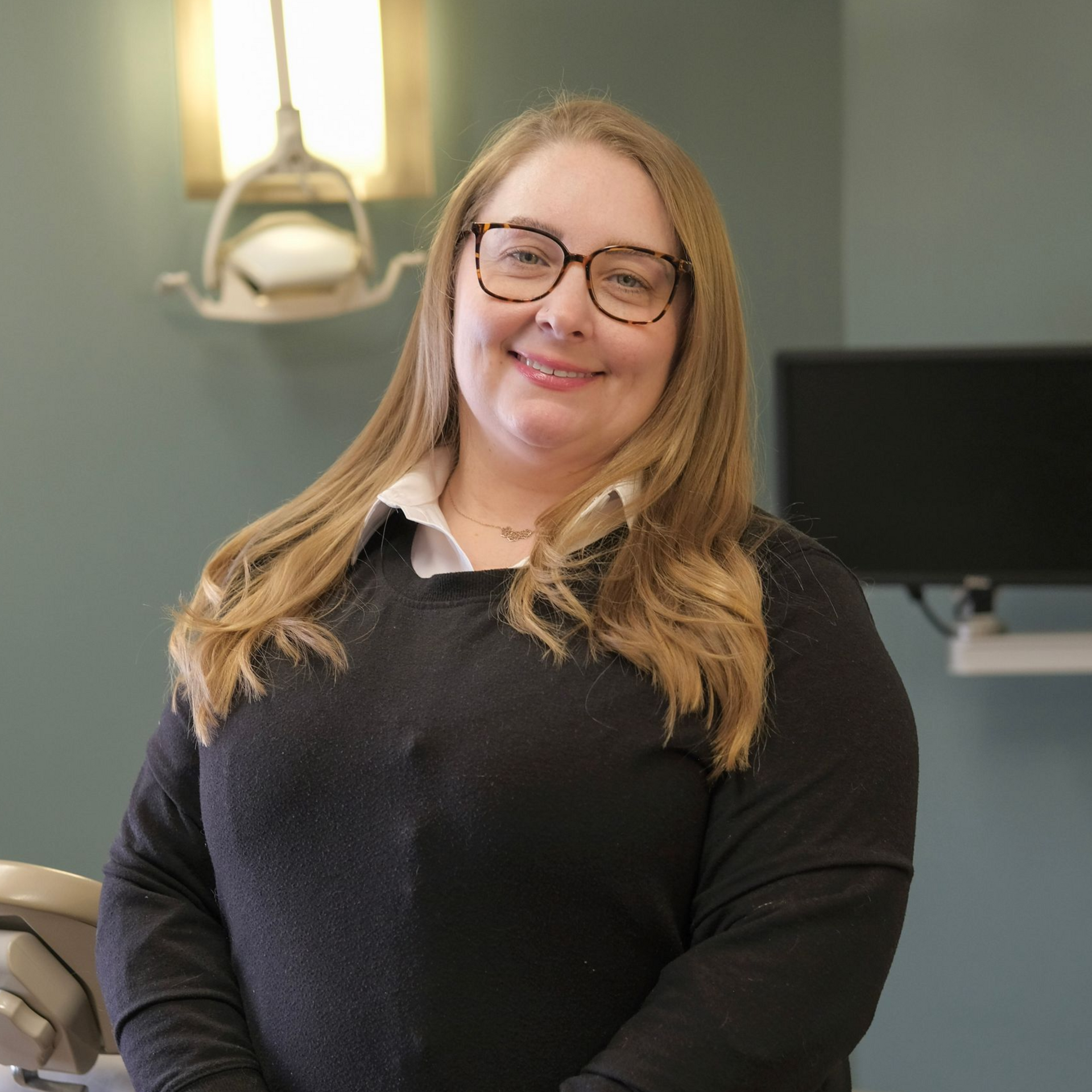 Sabrina with long auburn hair wearing glasses smiles at the camera against a blue background.