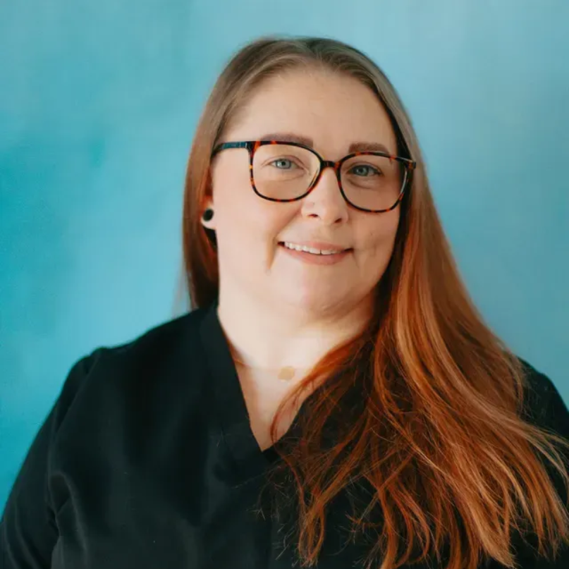 Sabrina with long auburn hair wearing glasses smiles at the camera against a blue background.