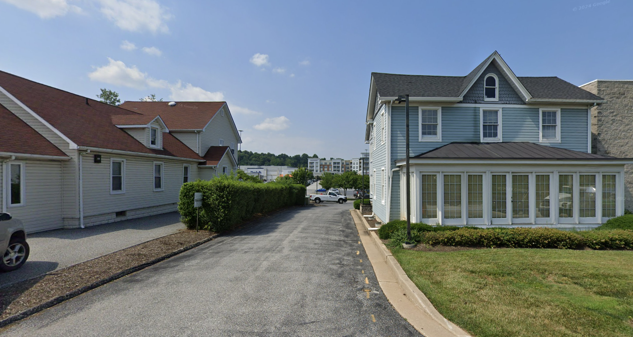 A paved driveway leads between a long white building on the left and a two-story blue house on the right under a blue sky.
