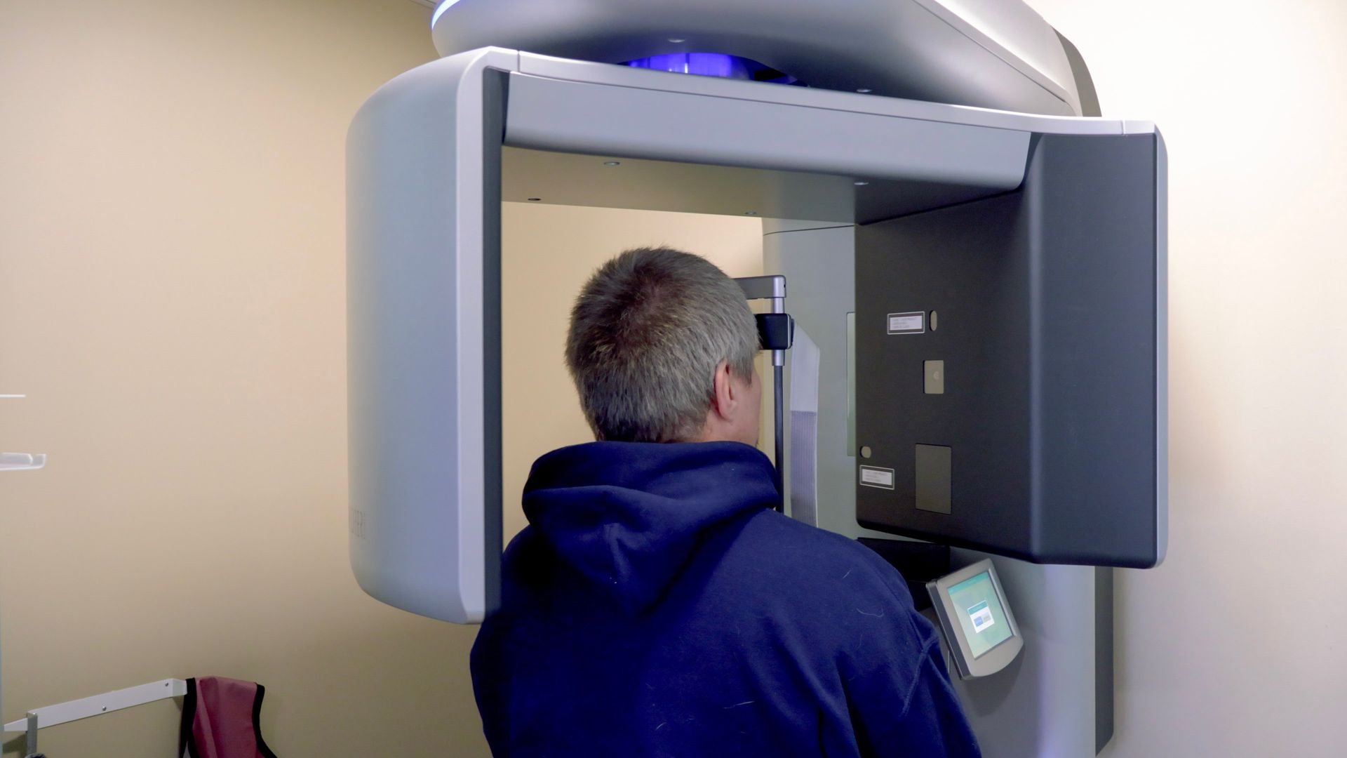A person in a blue hoodie standing with their head positioned inside a dental panoramic X-ray machine.