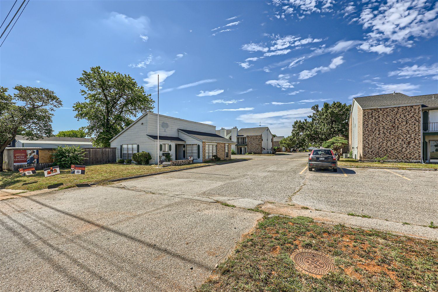 Check out our apartments for rent in Elk City, OK, at Crestview Commons.  This is an exterior view of an apartment building. The parking lot is visible, there are green trees in the background, and a bright blue sky with white clouds.