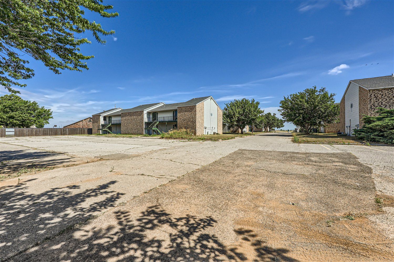 Check out our apartments for rent in Elk City, OK, at Crestview Commons.  This is an exterior view of an apartment building. The parking lot is visible, there are green trees in the background, and a bright blue sky with white clouds.