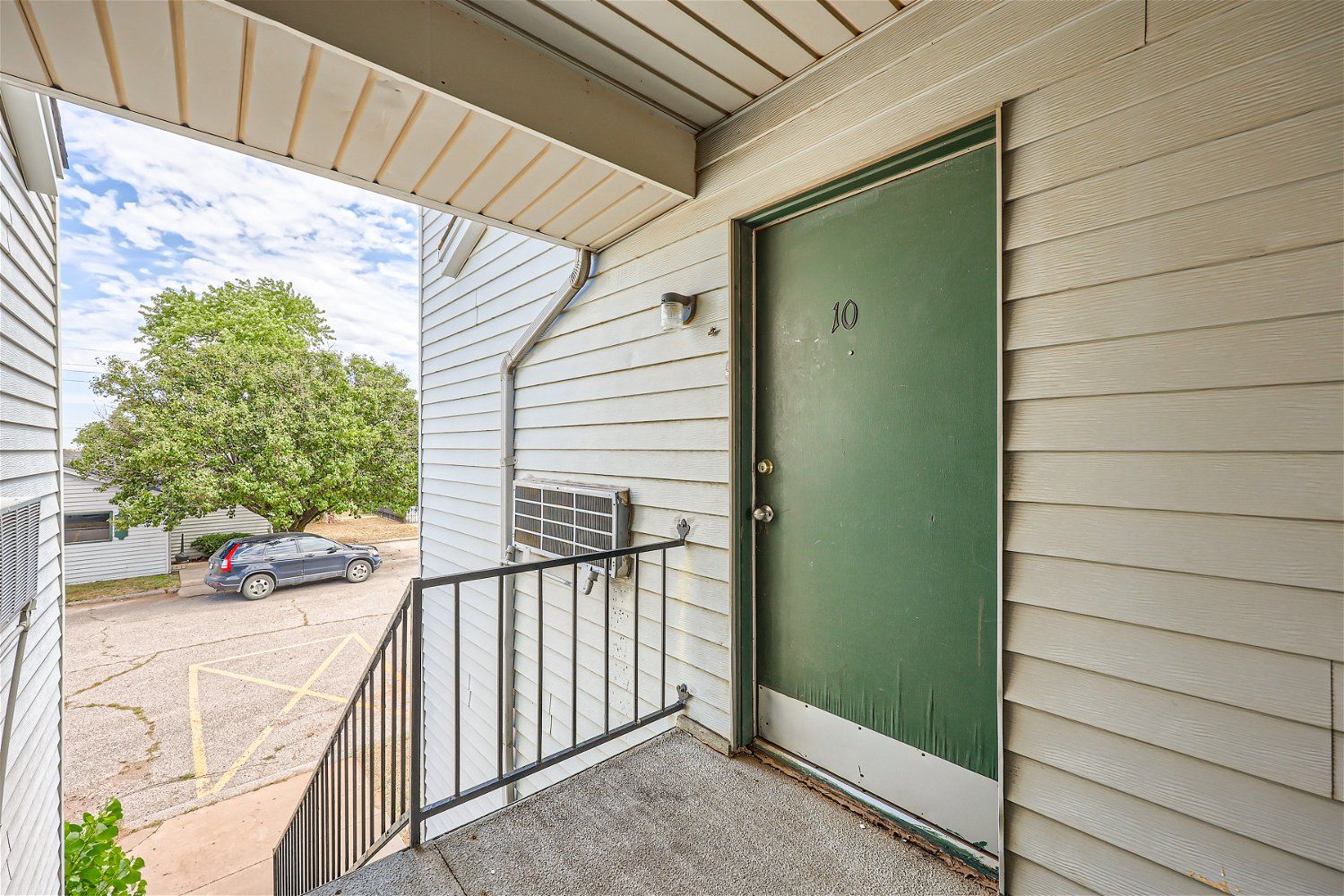 Apartment doorway with green door and small porch. Building exterior, car parked in lot.