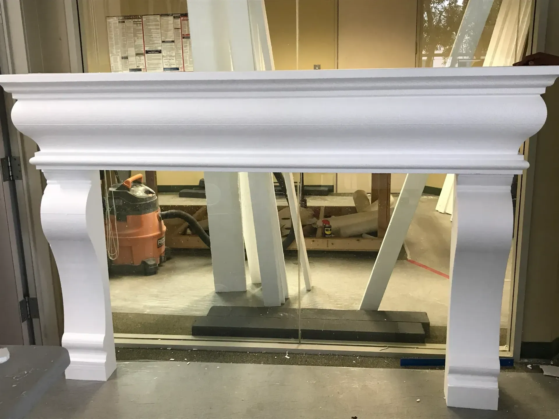 White painted fireplace mantel in a workshop, with tools and construction materials visible behind it.