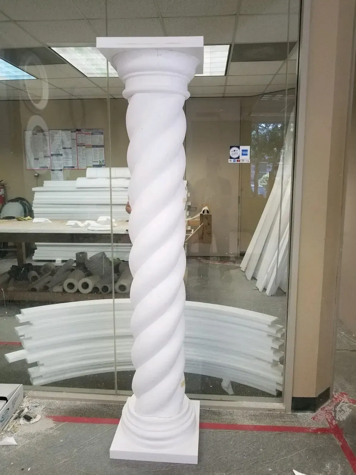 White twisted column standing in a storefront or workshop interior with glass walls and stacked materials behind it
