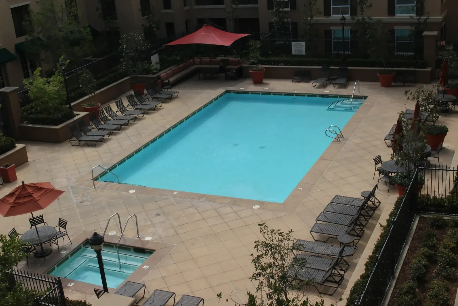 Outdoor hotel pool surrounded by lounge chairs and red umbrellas, with a small hot tub nearby.