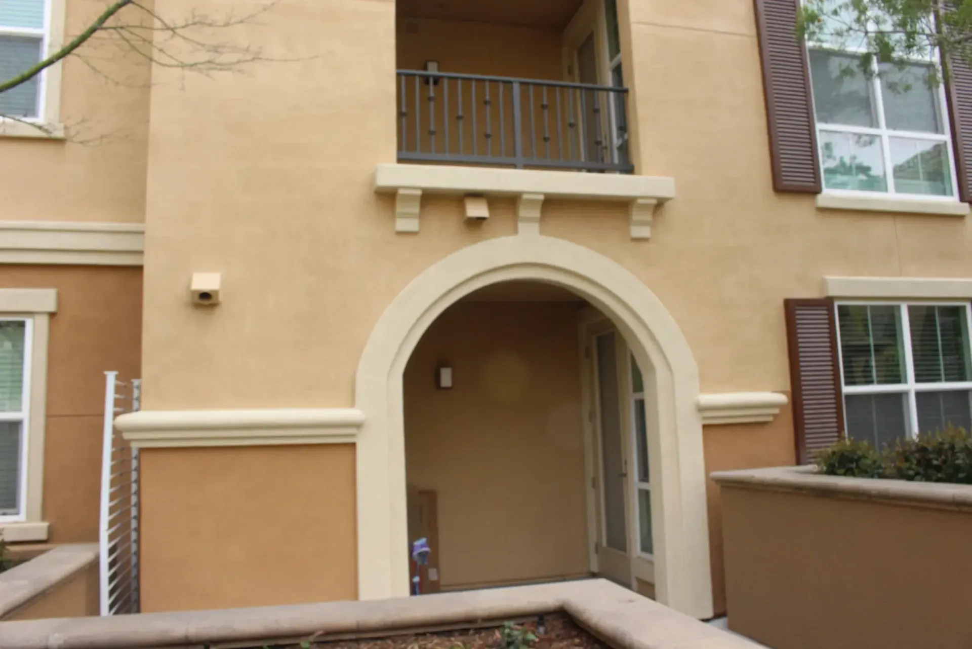 Tan stucco apartment entrance with arched doorway, balcony above, and windows with shutters on both sides