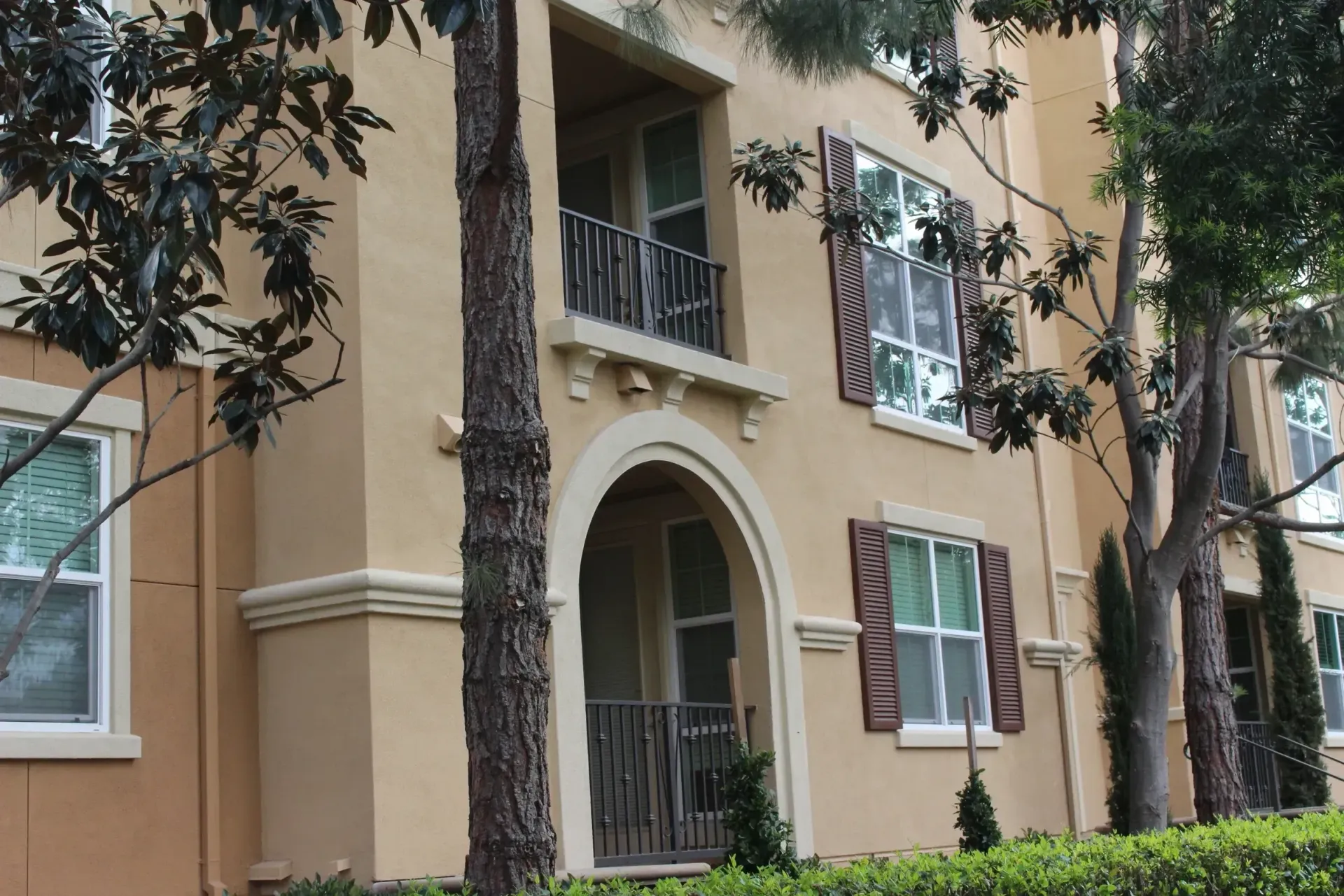 Tan stucco apartment building with arched entry, balconies, and trees in front