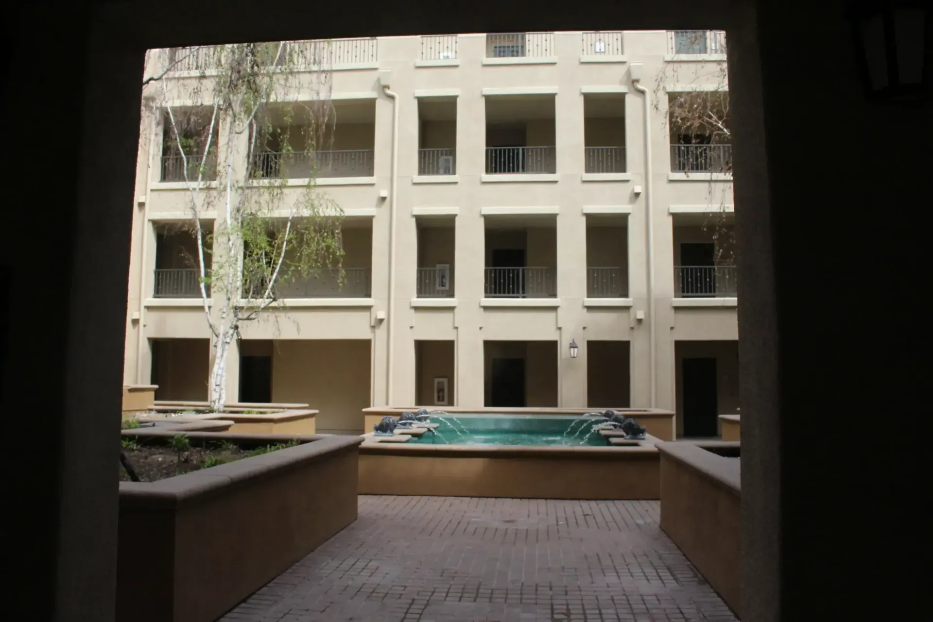 Courtyard view of an apartment building with balconies and trees, framed by a shaded walkway and planters