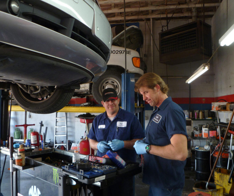 Two mechanics working on a car in a repair shop. One under a car, other looking at parts.