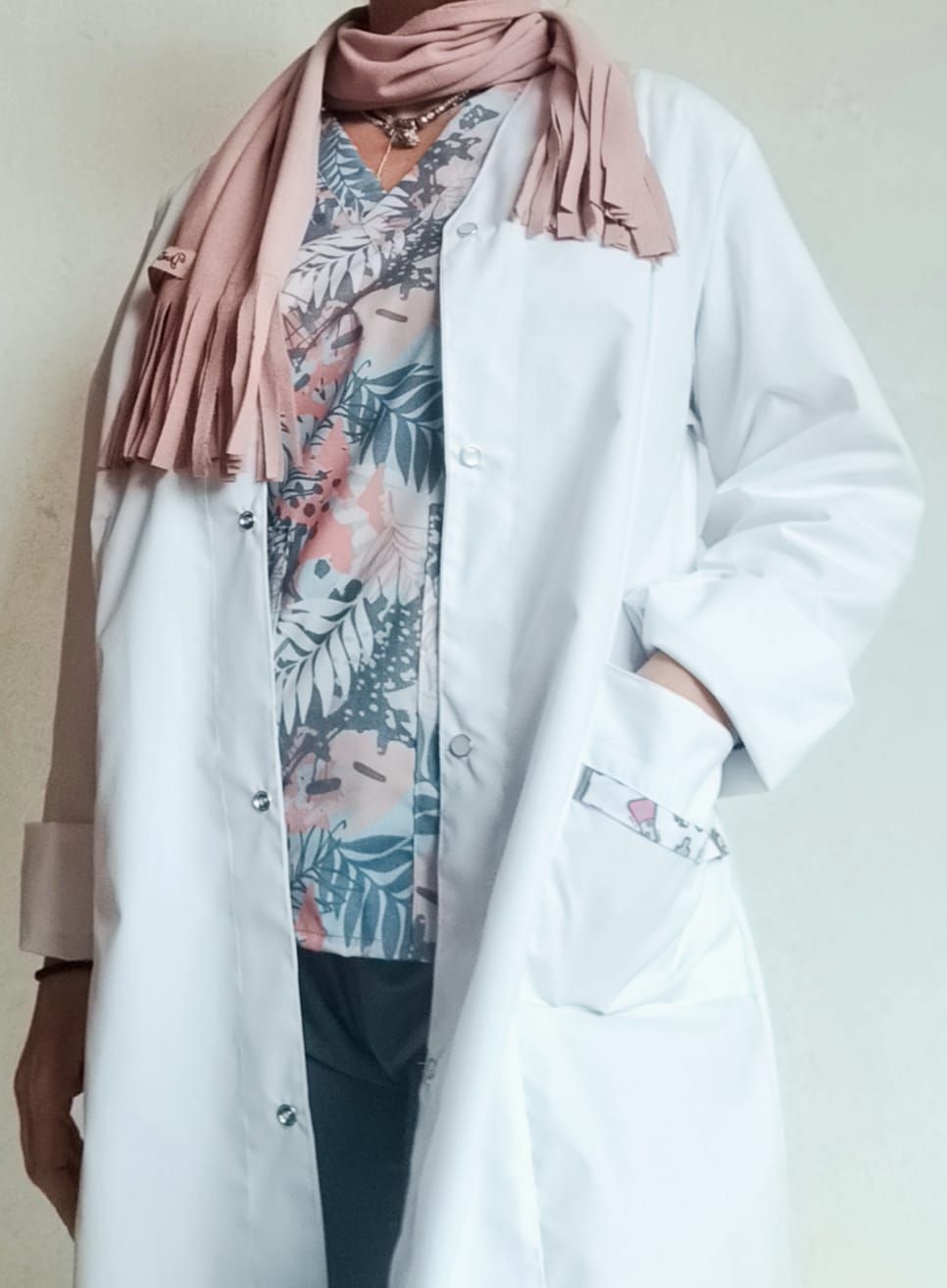 Persona con bata blanca, top floreado y bufanda rosa, con la mano en un bolsillo decorado con el mismo estampado floral.