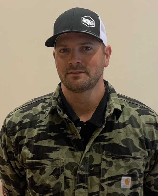 A man wearing a hat and a camouflage shirt is standing in front of a wall.