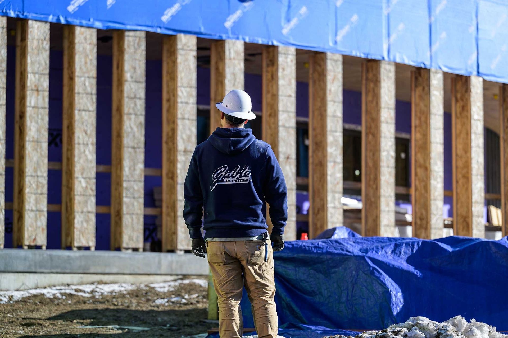 Construction worker wearing a hard hat, facing a building under construction; wood framing, blue tarp.