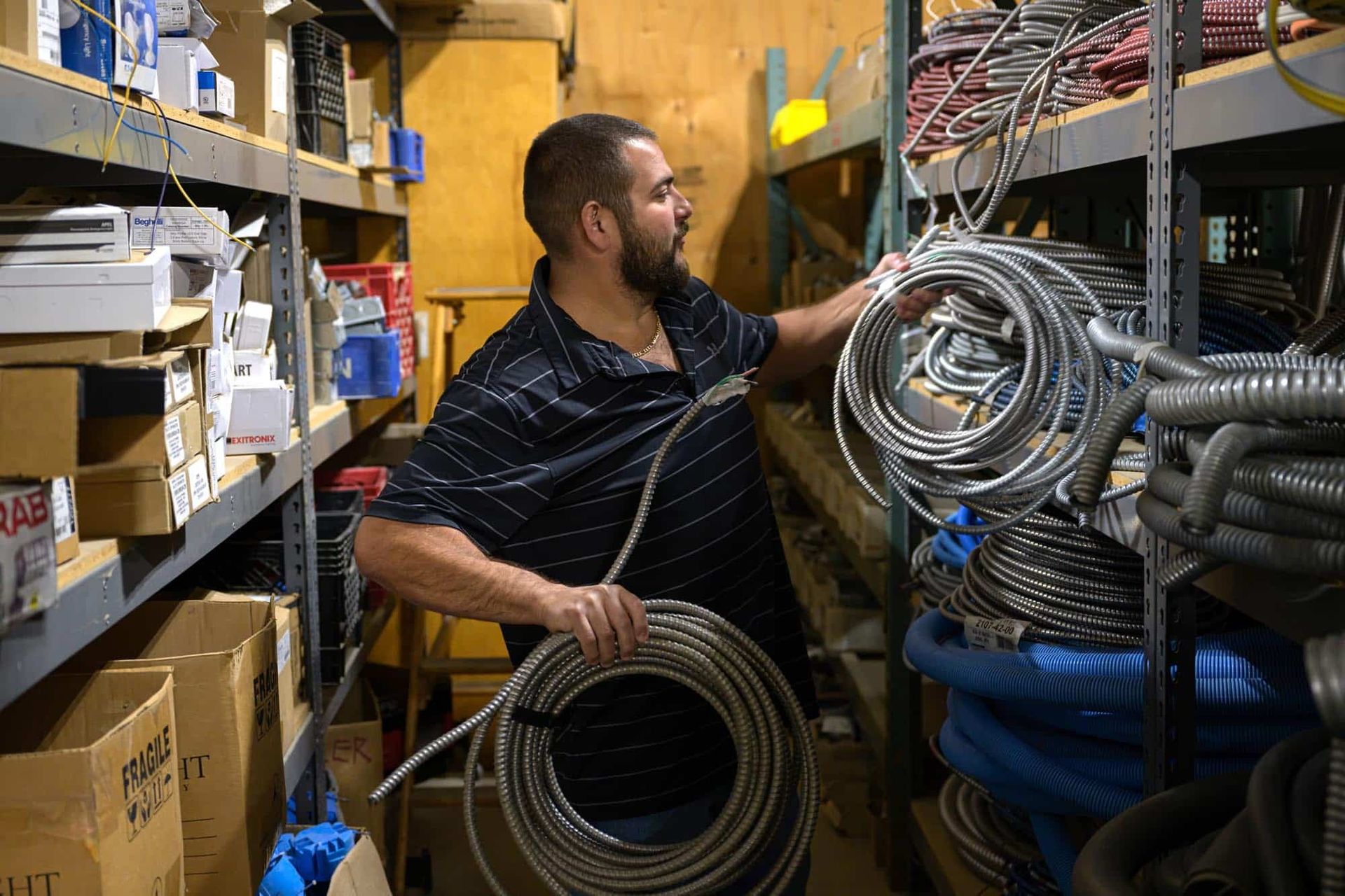 Man in a warehouse holding a coiled metal hose; surrounded by shelves of parts.