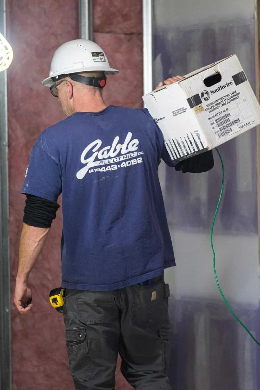 Construction worker holding wire box, in a partially built wall. Blue shirt, hard hat, and gray pants.