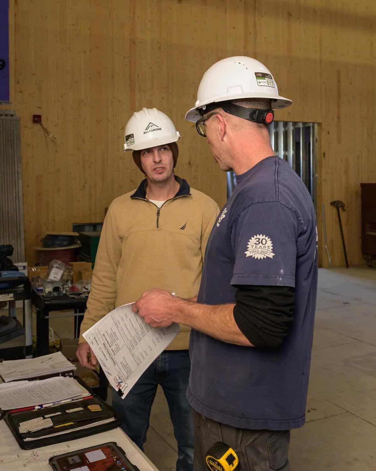 Two men wearing hard hats examine papers in a construction setting. One points to the document.