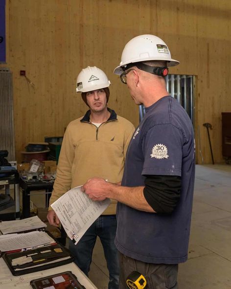 Two men wearing hard hats examine papers in a construction setting. One points to the document.