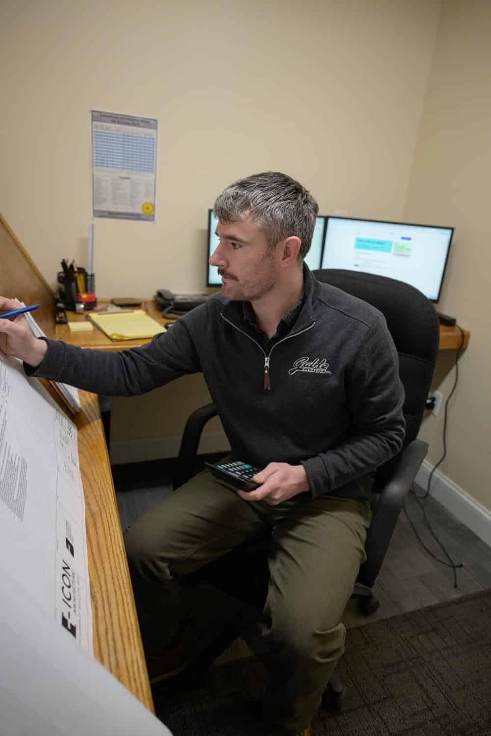 Man with gray hair and beard working on drawings at a desk with dual monitors.