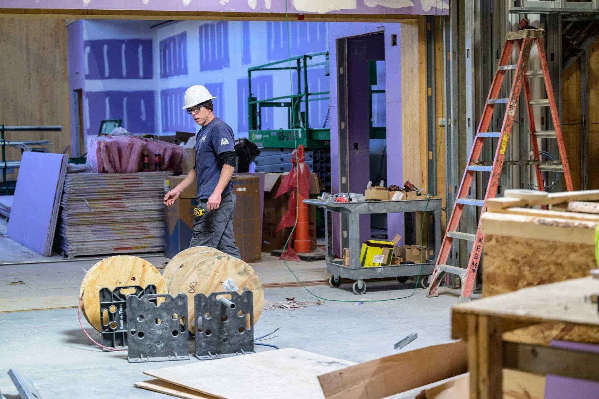 Construction worker in hard hat walks through a room. Purple walls, tools, and a ladder are visible.