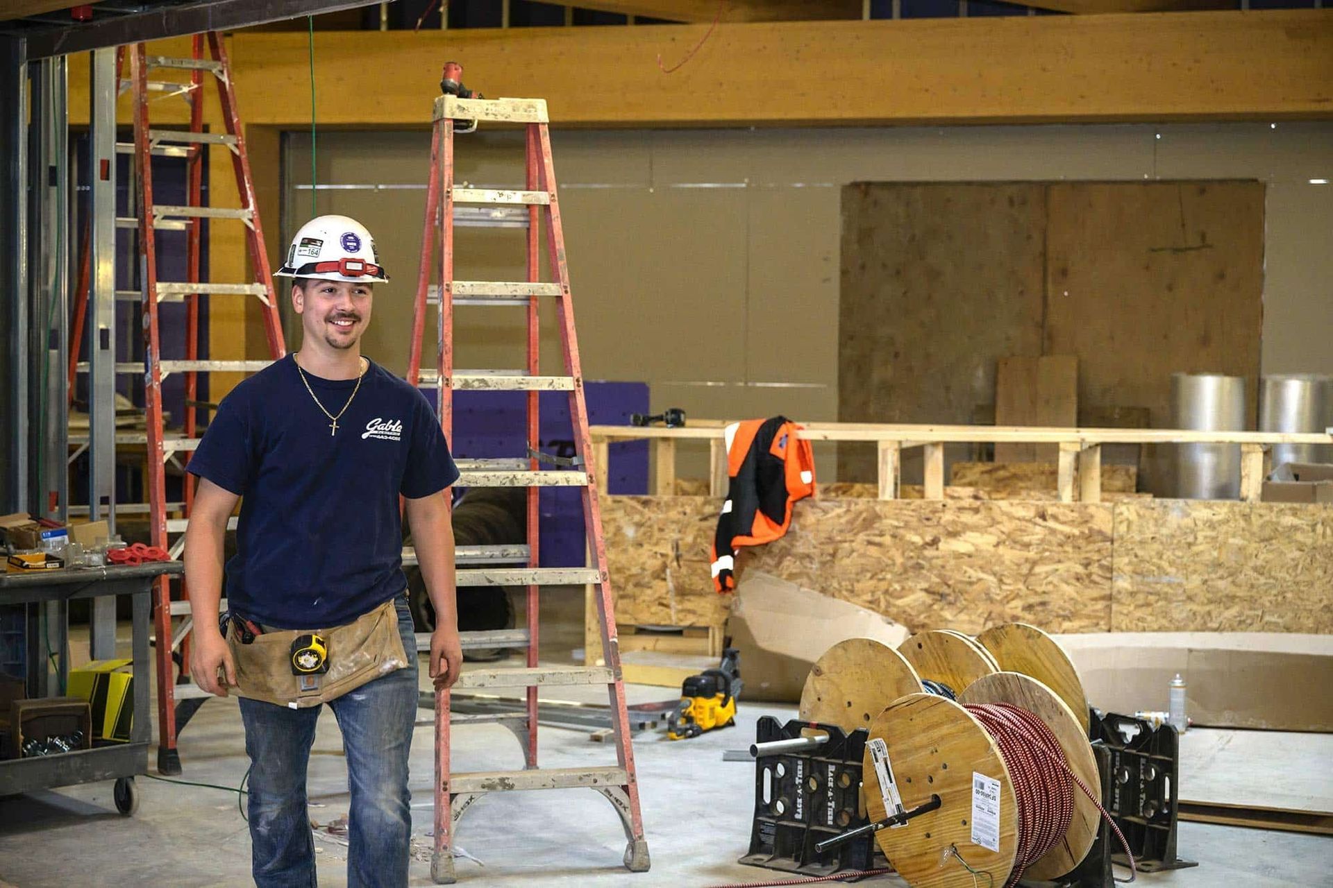 Construction worker stands in a partially built room, wearing a hard hat and tool belt, with ladders and spools of cable.
