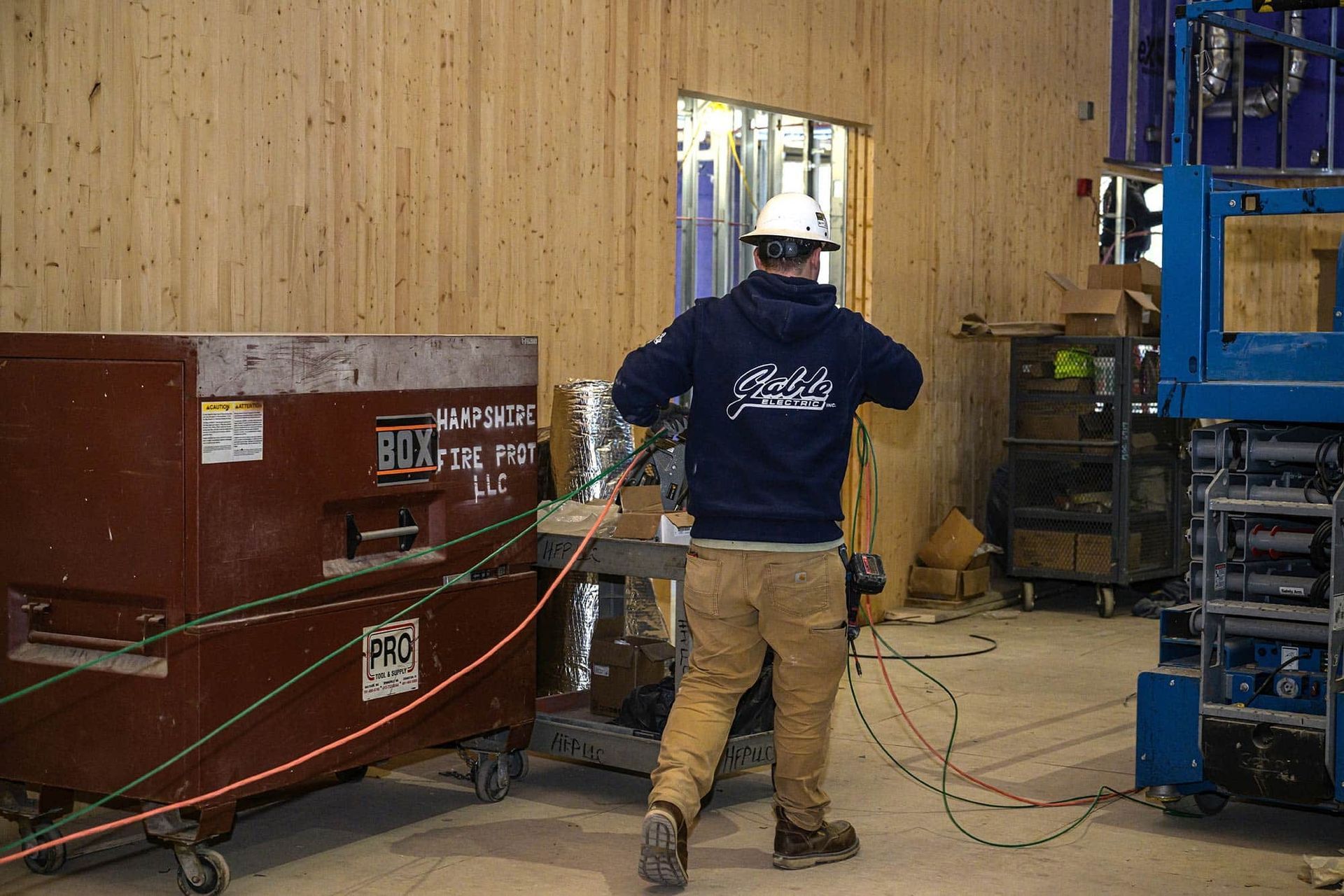 Construction worker in hard hat drilling near dumpster, wires, and equipment in a wood-paneled room.