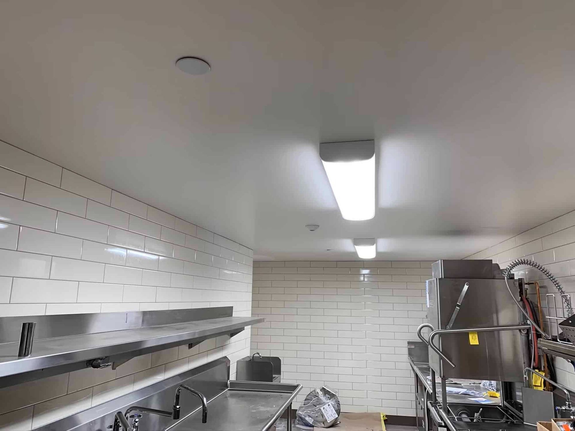 Commercial kitchen with stainless steel appliances, white tile walls, and fluorescent lighting.