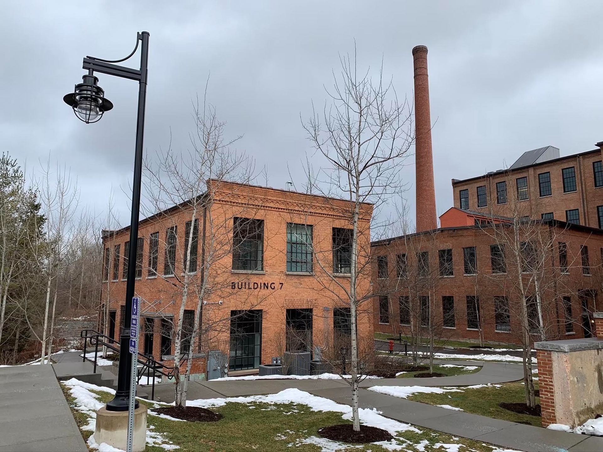 Brick buildings with a smokestack and lamp post, snowy ground, overcast sky.