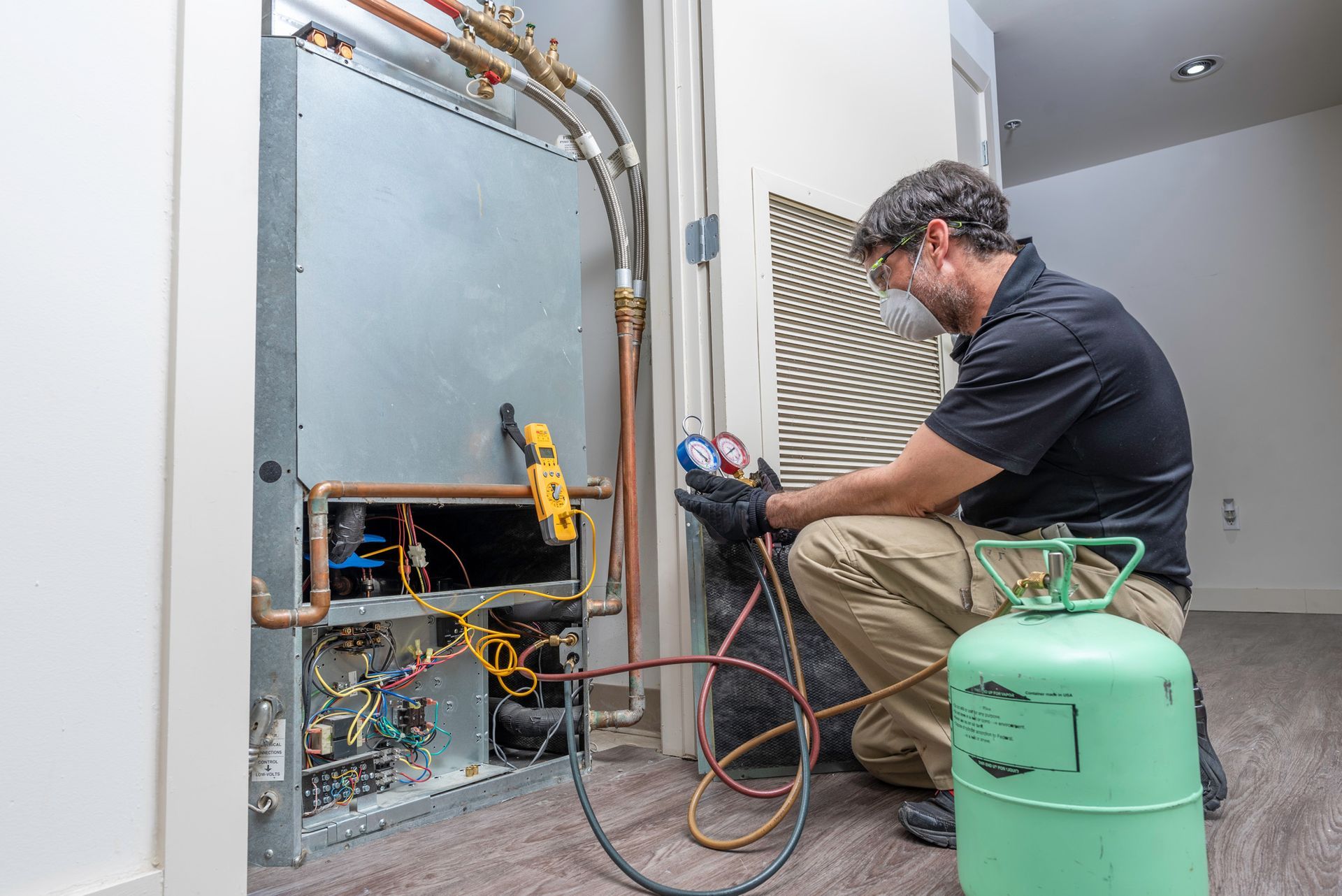 HVAC technician working on an air conditioning unit, checking gauges. Indoors with equipment and a green tank.