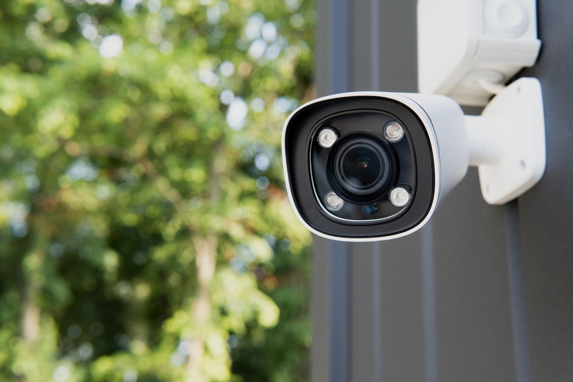 White security camera mounted on a dark wall, focused on the viewer with a green tree background.