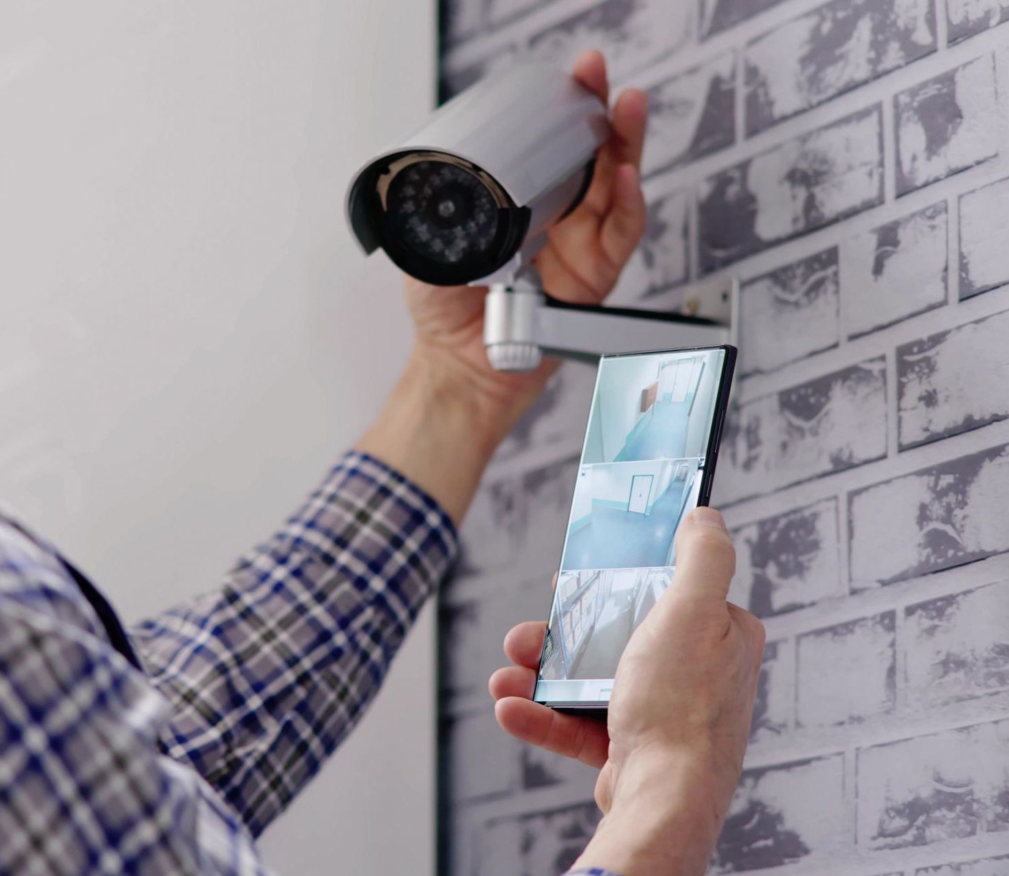 Person installing a security camera, looking at video feed on a smartphone against a brick wall.
