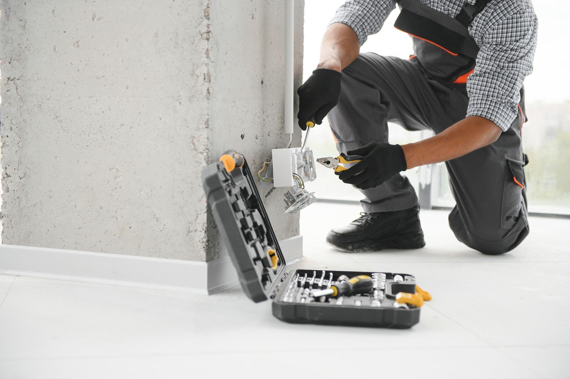Electrician in work clothes installing an electrical outlet, tools in open case nearby.