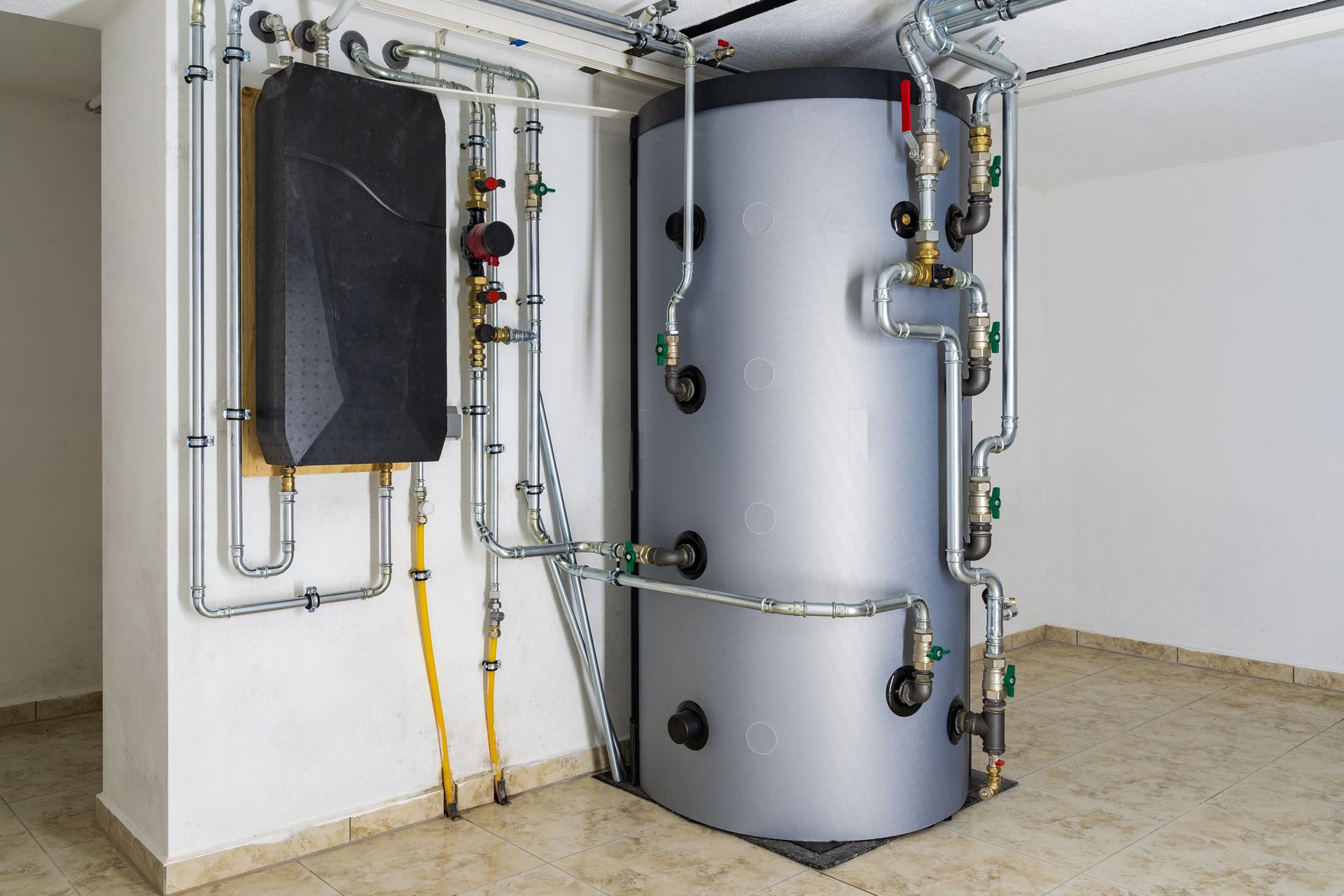 Water heater and auxiliary tank with connected pipes in a utility room, against a white wall.