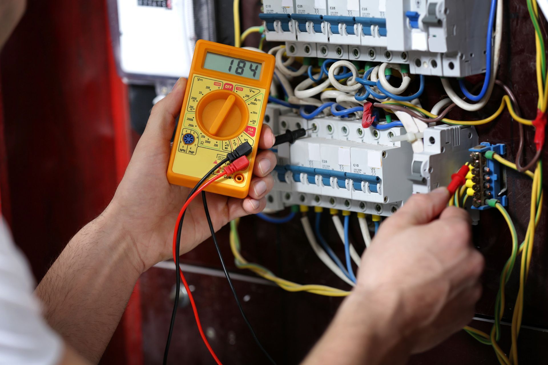 Person using a multimeter to test electrical panel wiring; digital display reads 184 volts.