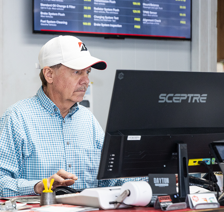 Man at desk with computer, white hat, blue plaid shirt; auto shop setting, looking at screen | Dayton Tire & Auto