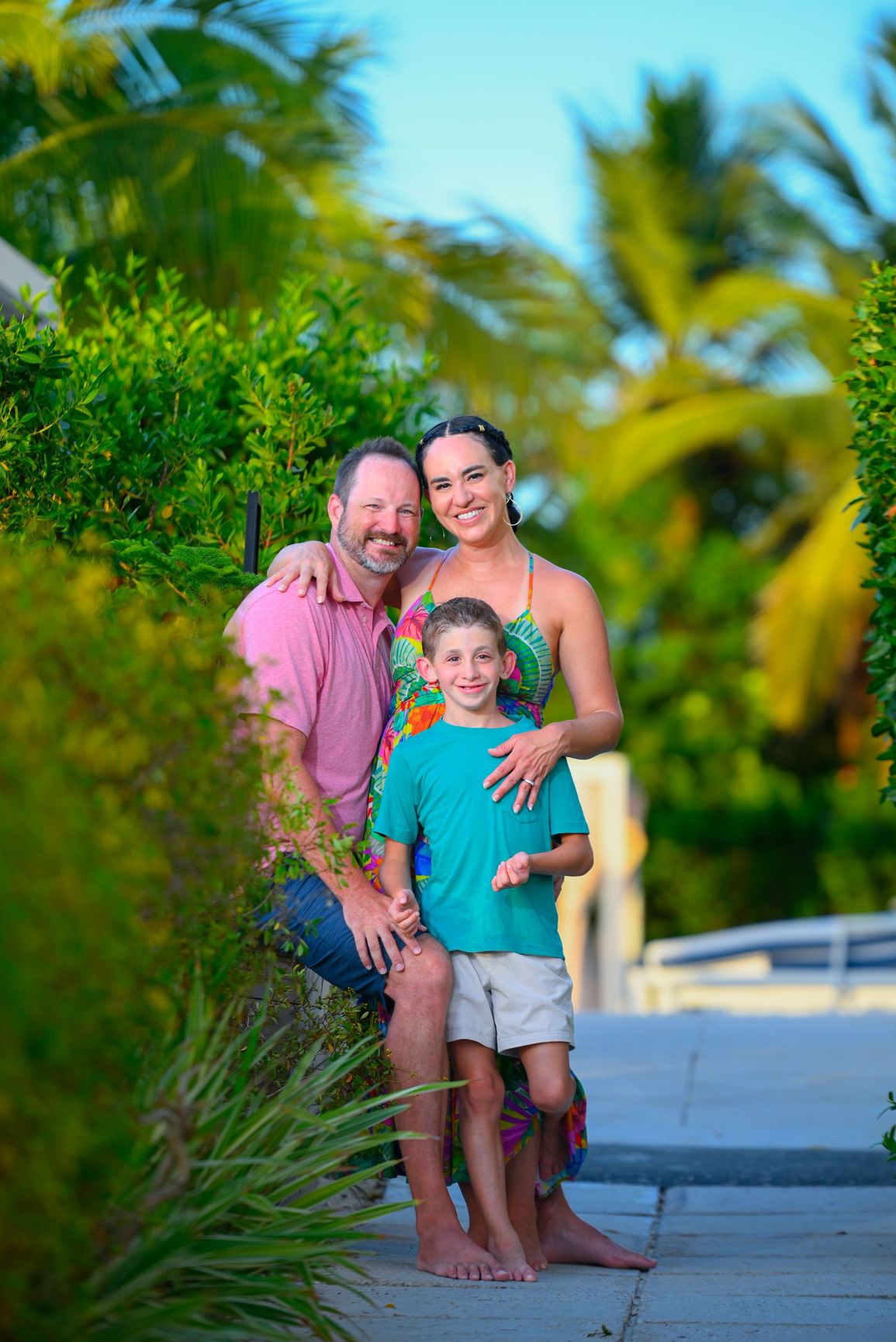 Family of three poses by a tree. Woman in white dress, man in blue shirt, child in blue on branch.