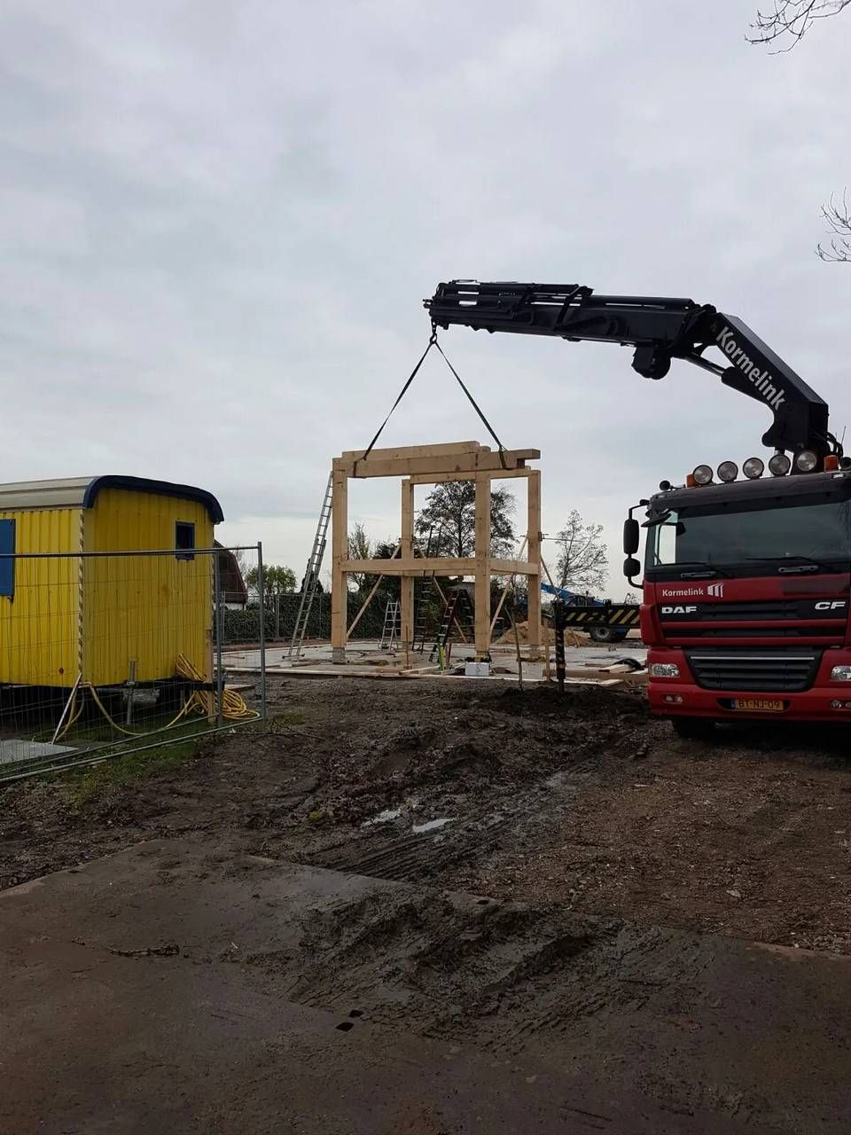 Een rode vrachtwagen met een kraan erop draagt een houten constructie.